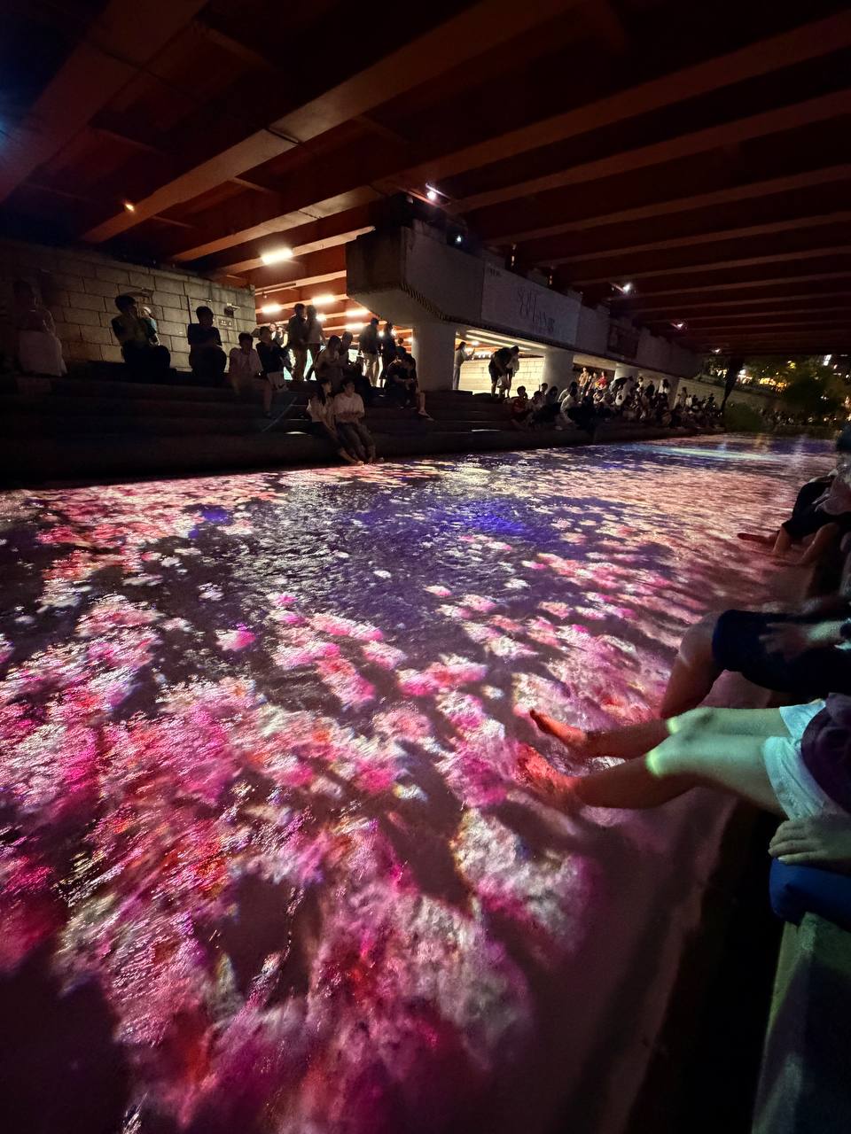 Cheonggyecheon waterfall lit with pink and purple light projections people dipping feet in water Seoul